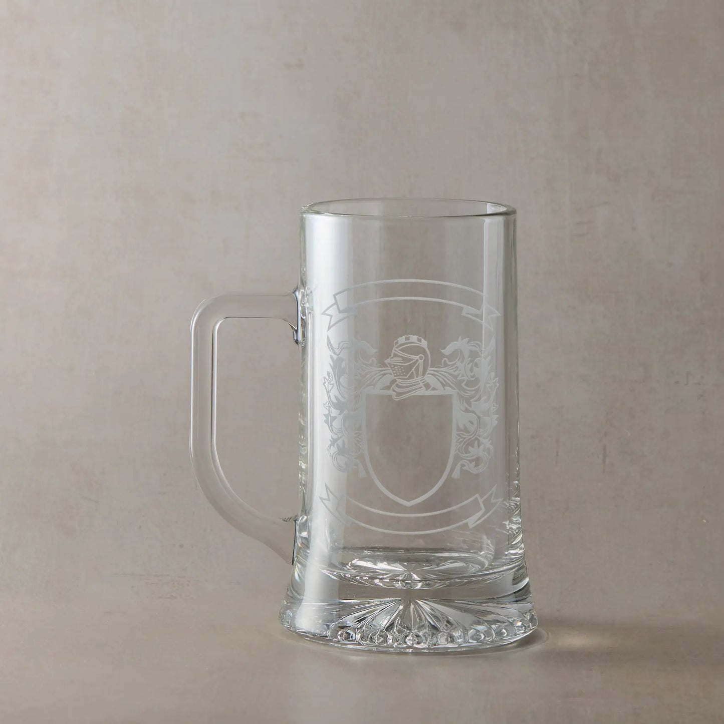 Engraved glass tankard featuring a heraldic coat of arms with a central shield left clear for your own design, handle on the left, set against a neutral beige studio background