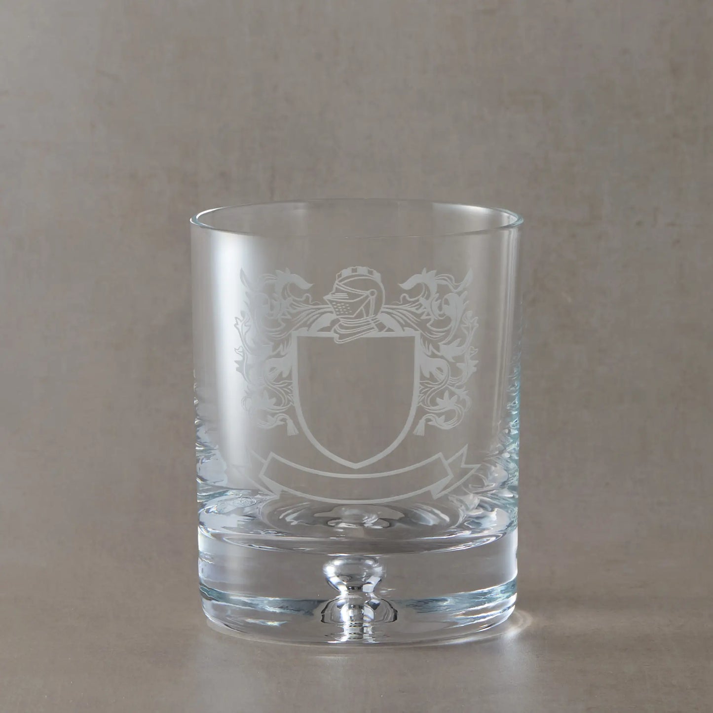 Engraved whisky glass with heraldic coat of arms design on a bubble base tumbler, shown on a neutral beige studio background