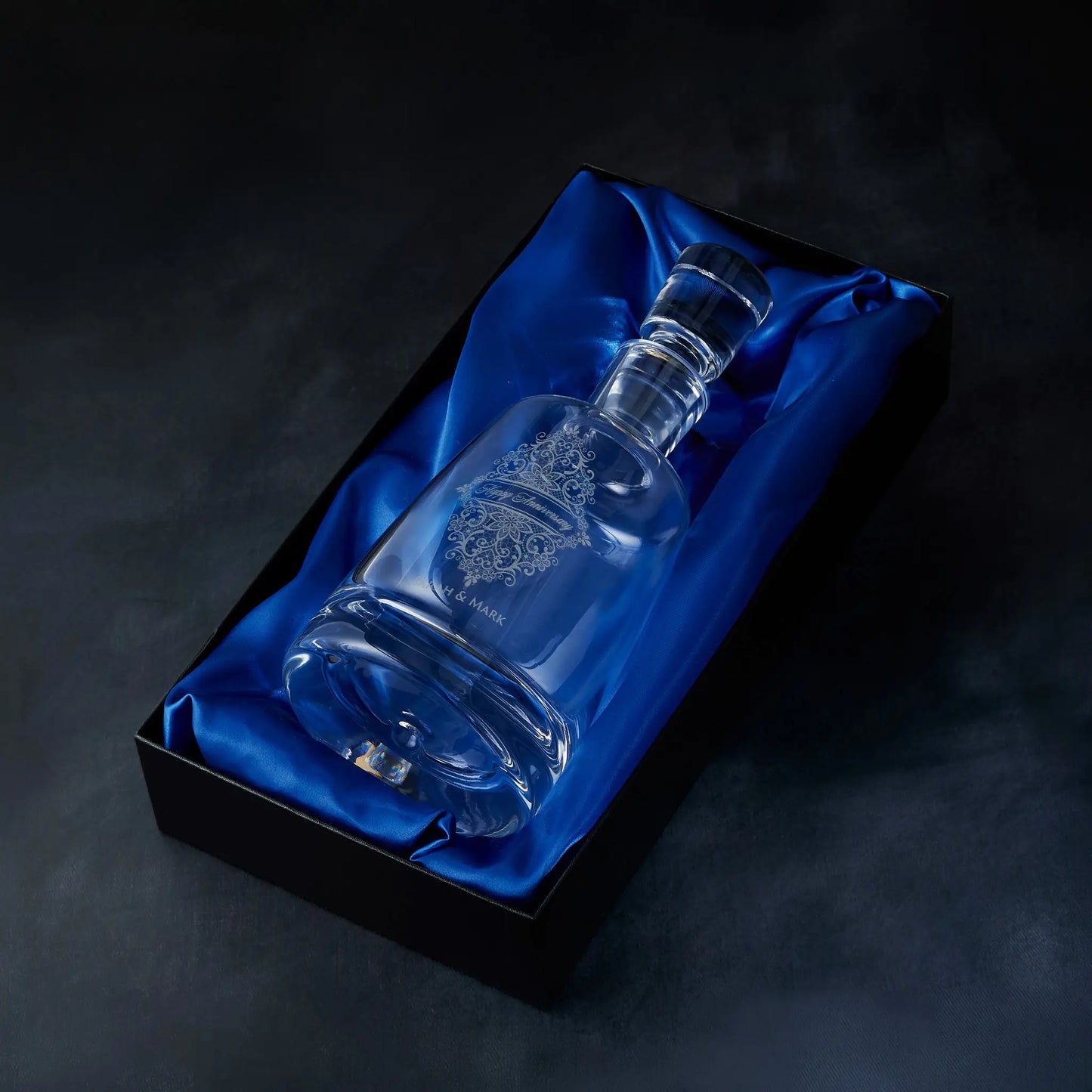 Engraved guipure lace spirit decanter presented in a satin-lined gift box, photographed against a deep blue background.