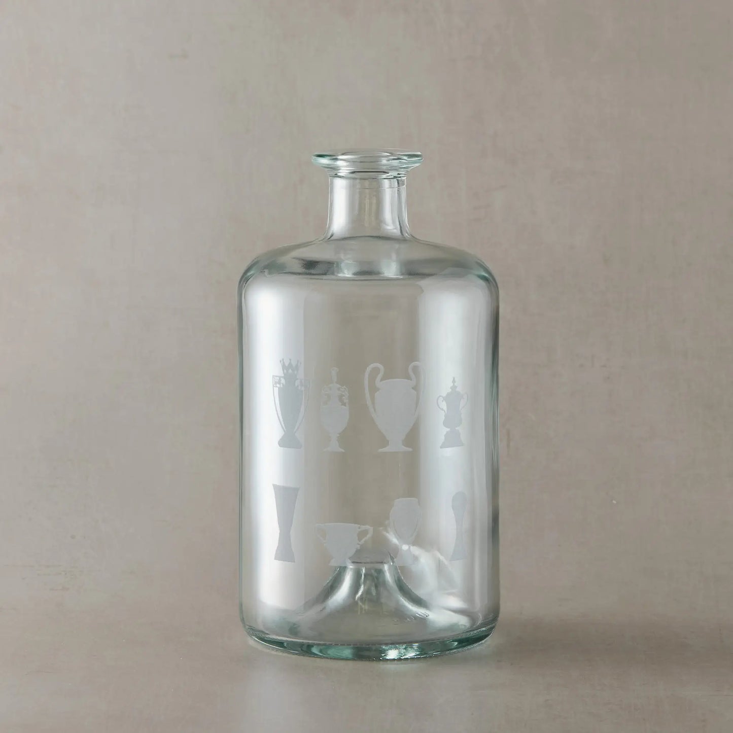 Apothecary-style glass decanter engraved with eight trophy silhouettes photographed on a beige background