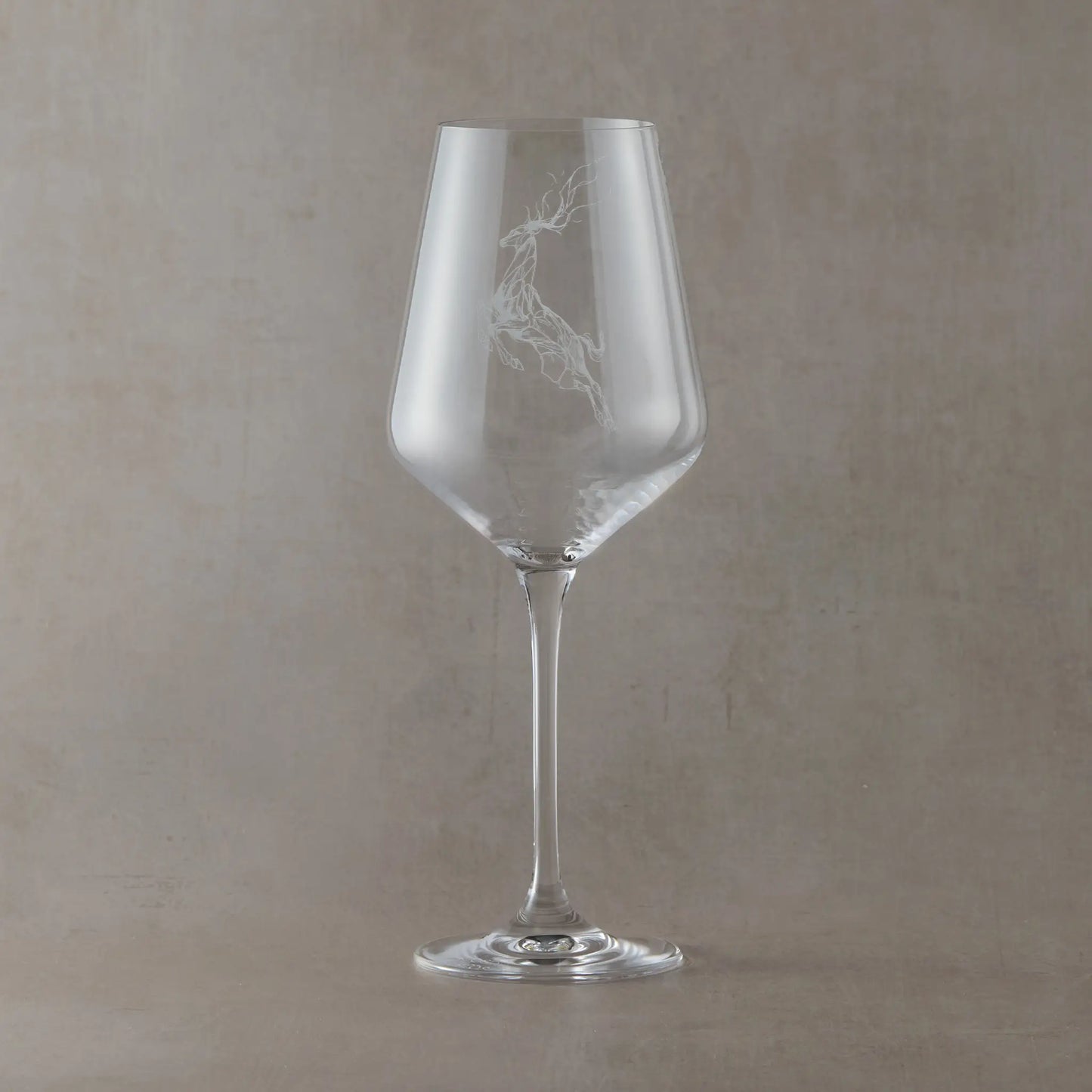 Clear wine glass engraved with a stag and scattered stars on a soft neutral beige background