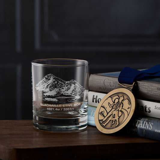 whisky glass with Buachaille Etive Mor mountain engraving, books, and a hillwalking medal on a wooden surface
