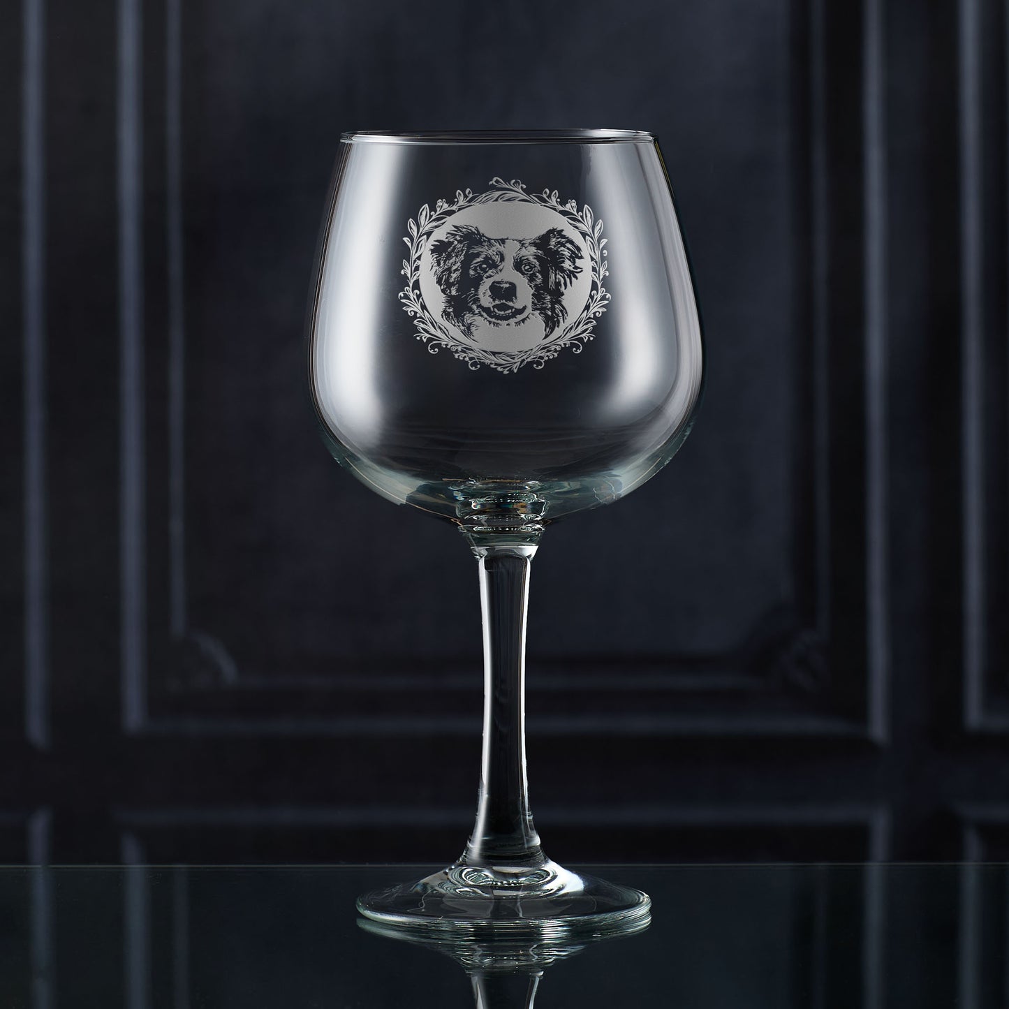 Hand-engraved Border Collie gin glass shown in front of a dark panelled wall, featuring a leafy garland and detailed Collie portrait design by Just The Ticket.