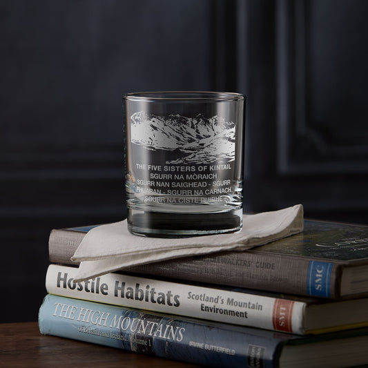 Glass whisky tumbler with the Five Sisters of Kintail and engraved text on a stack of books against a dark background