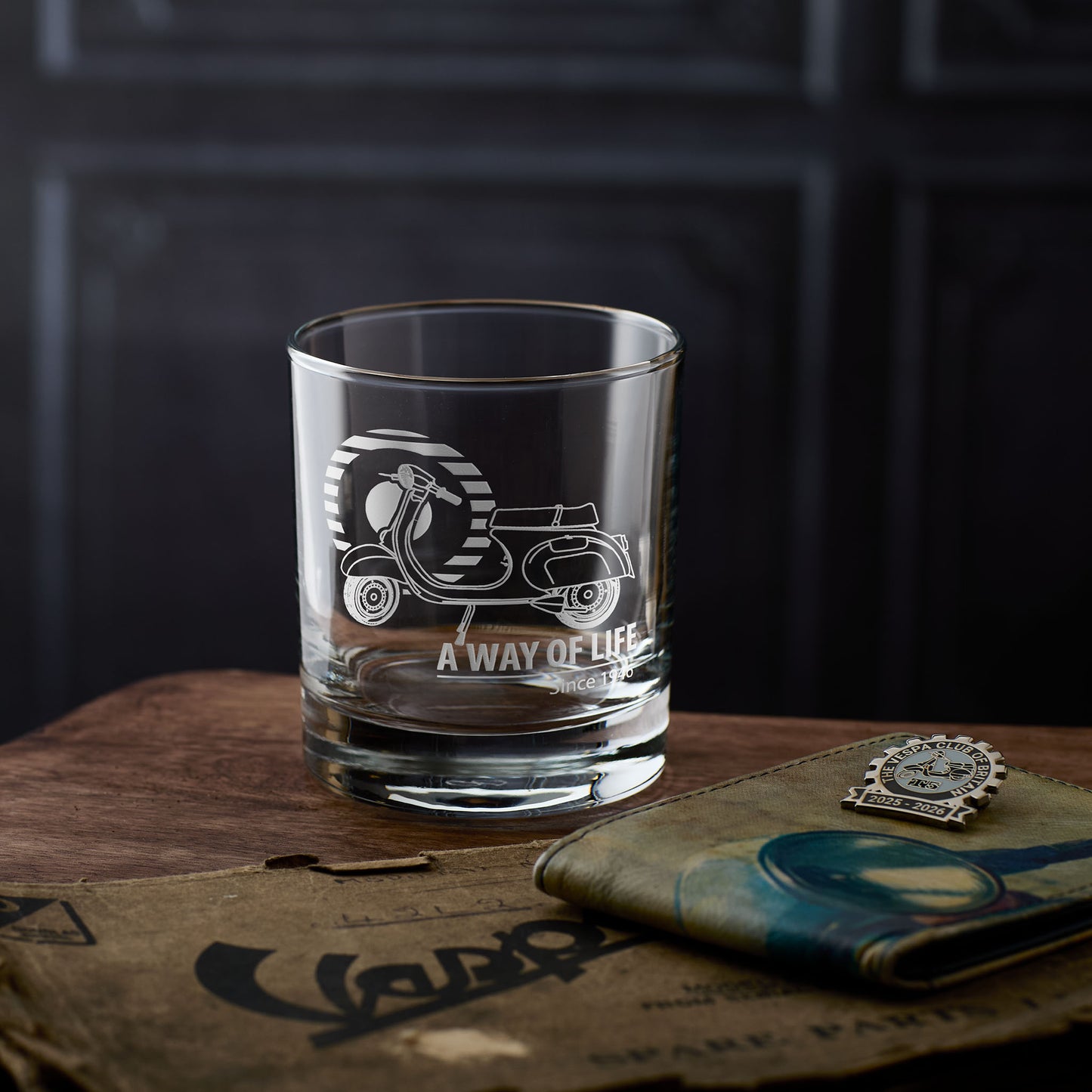 Whisky glass with engraved Vespa Primavera design and text on a wooden surface with a dark background