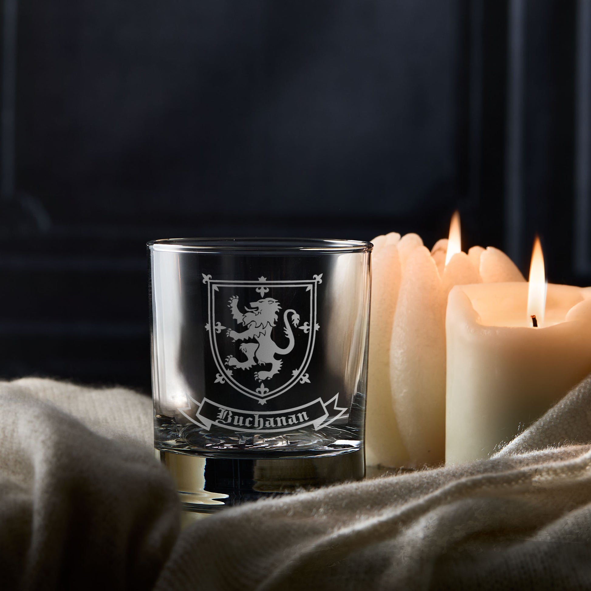 Coat of Arms whisky glass with engraved design and 'Buchanan' family crest, surrounded by candles on a dark background