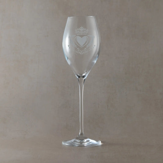Clear prosecco glass engraved with a Claddagh heart, crown and hands motif, photographed on a soft neutral beige background