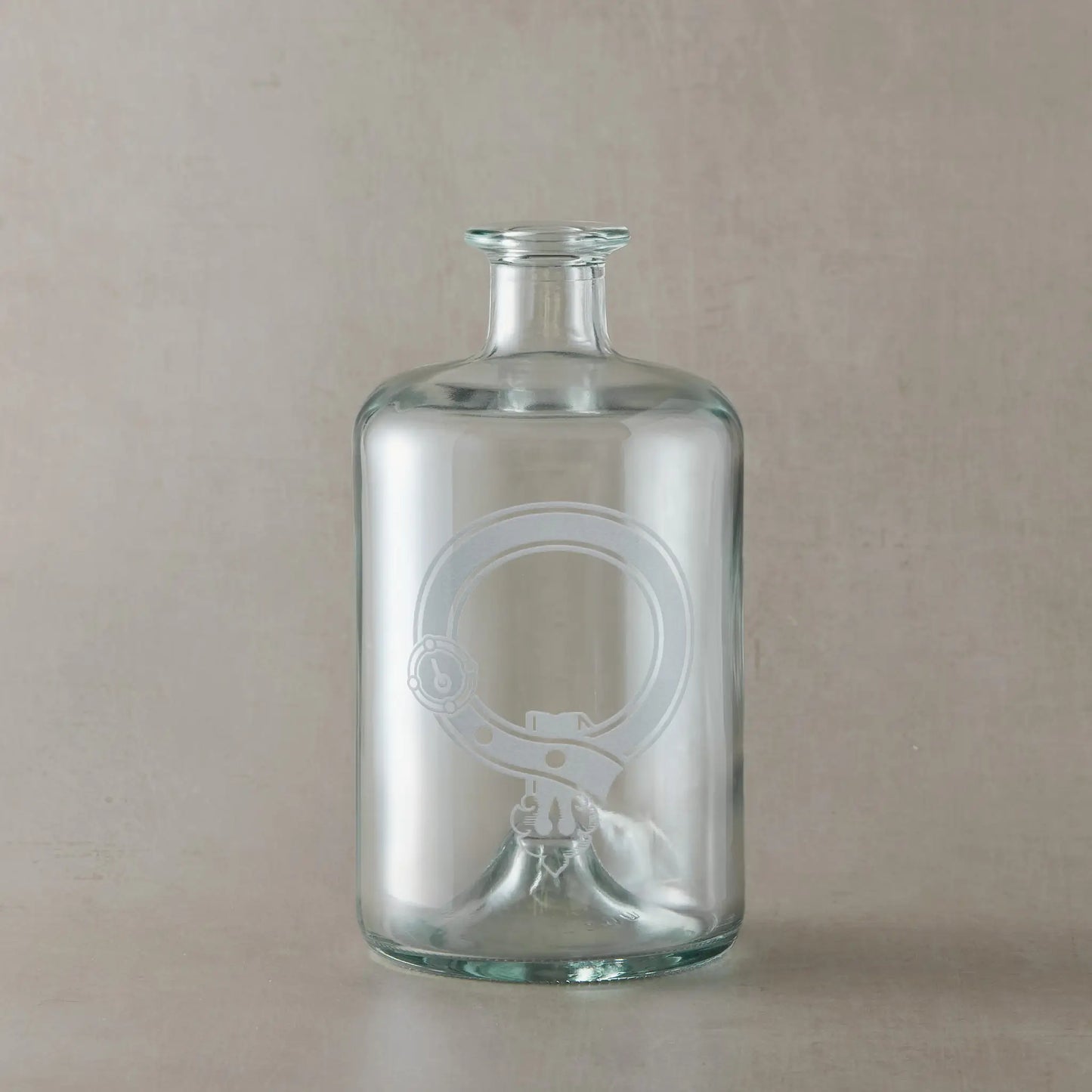 Engraved apothecary decanter featuring a Scottish clan crest with belt and buckle motif, centre left blank for your own design, set on a clear glass bottle against a neutral beige studio background