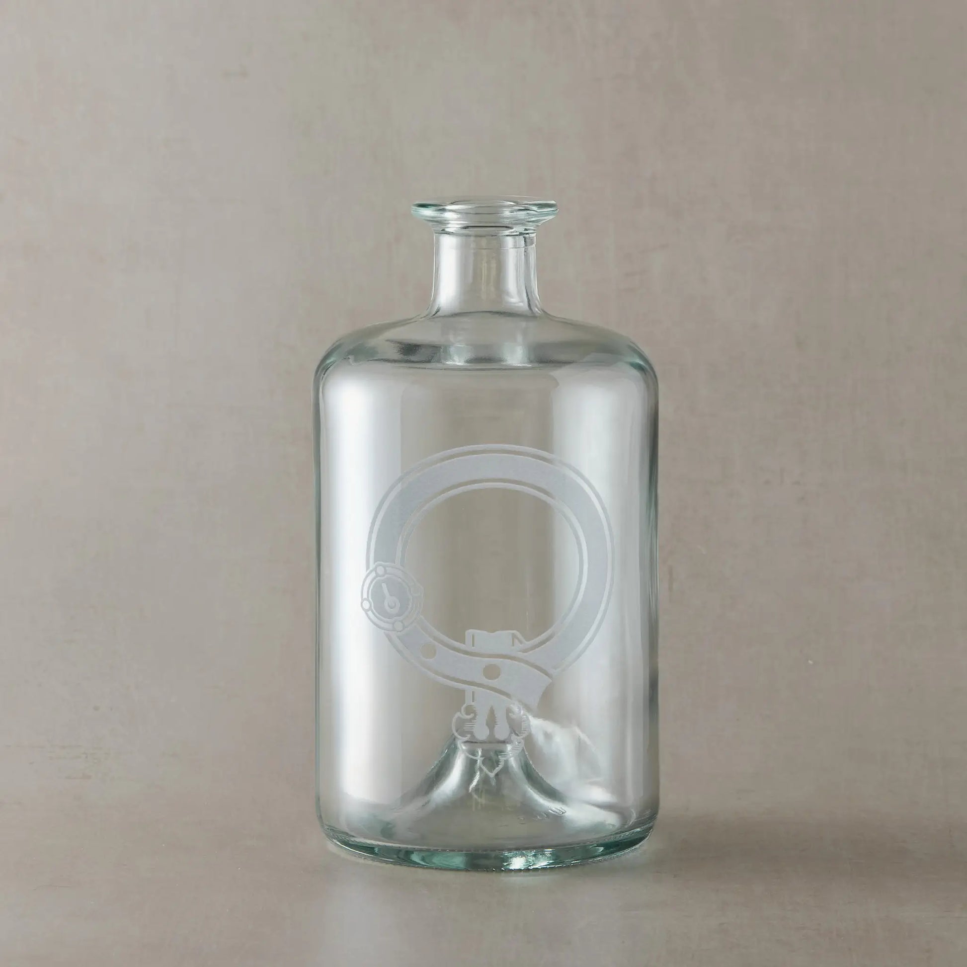 Engraved apothecary decanter featuring a Scottish clan crest with belt and buckle motif, centre left blank for your own design, set on a clear glass bottle against a neutral beige studio background