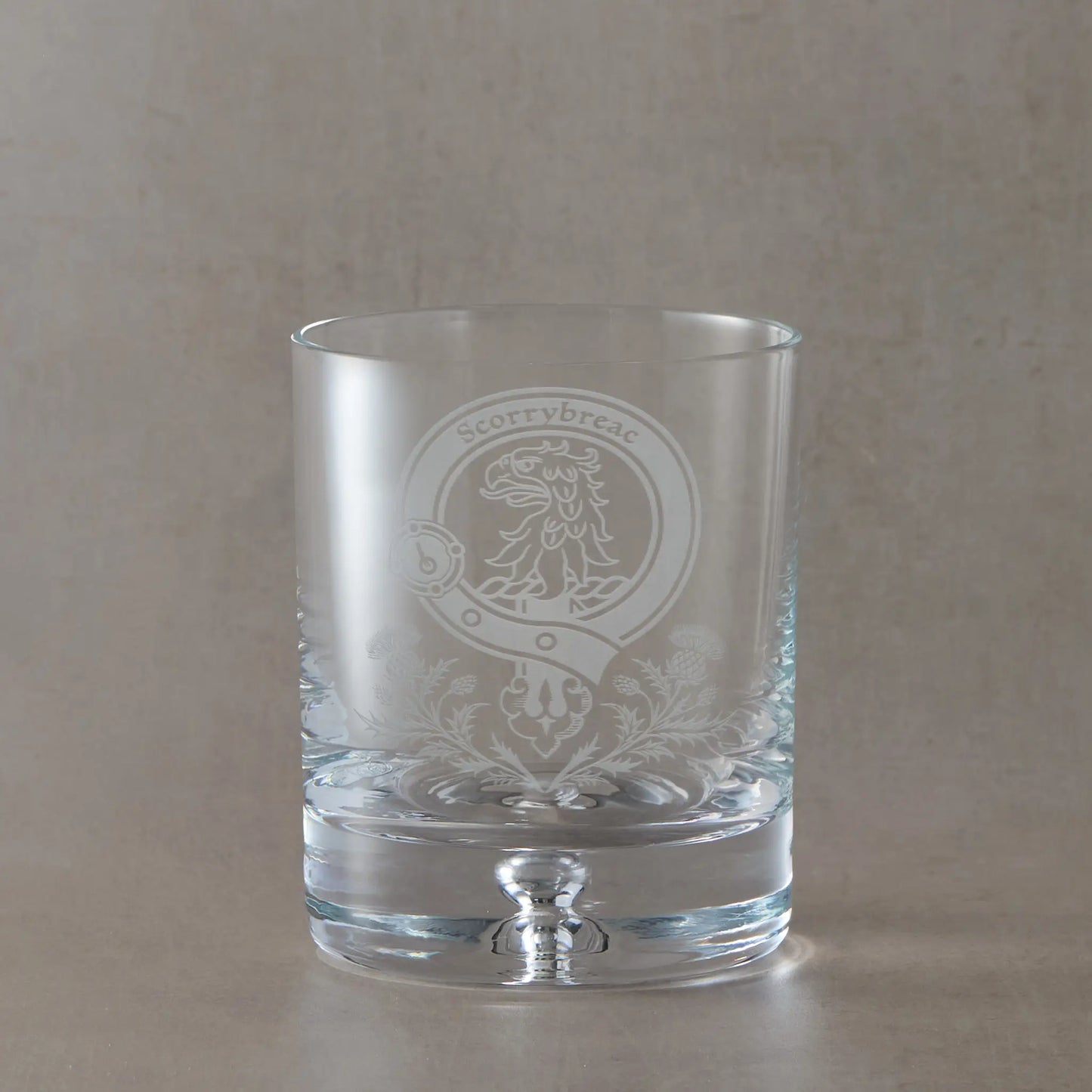 Engraved whisky glass featuring a Scottish clan crest with belt and buckle motif, sandblasted on a clear glass tumbler against a neutral beige studio background