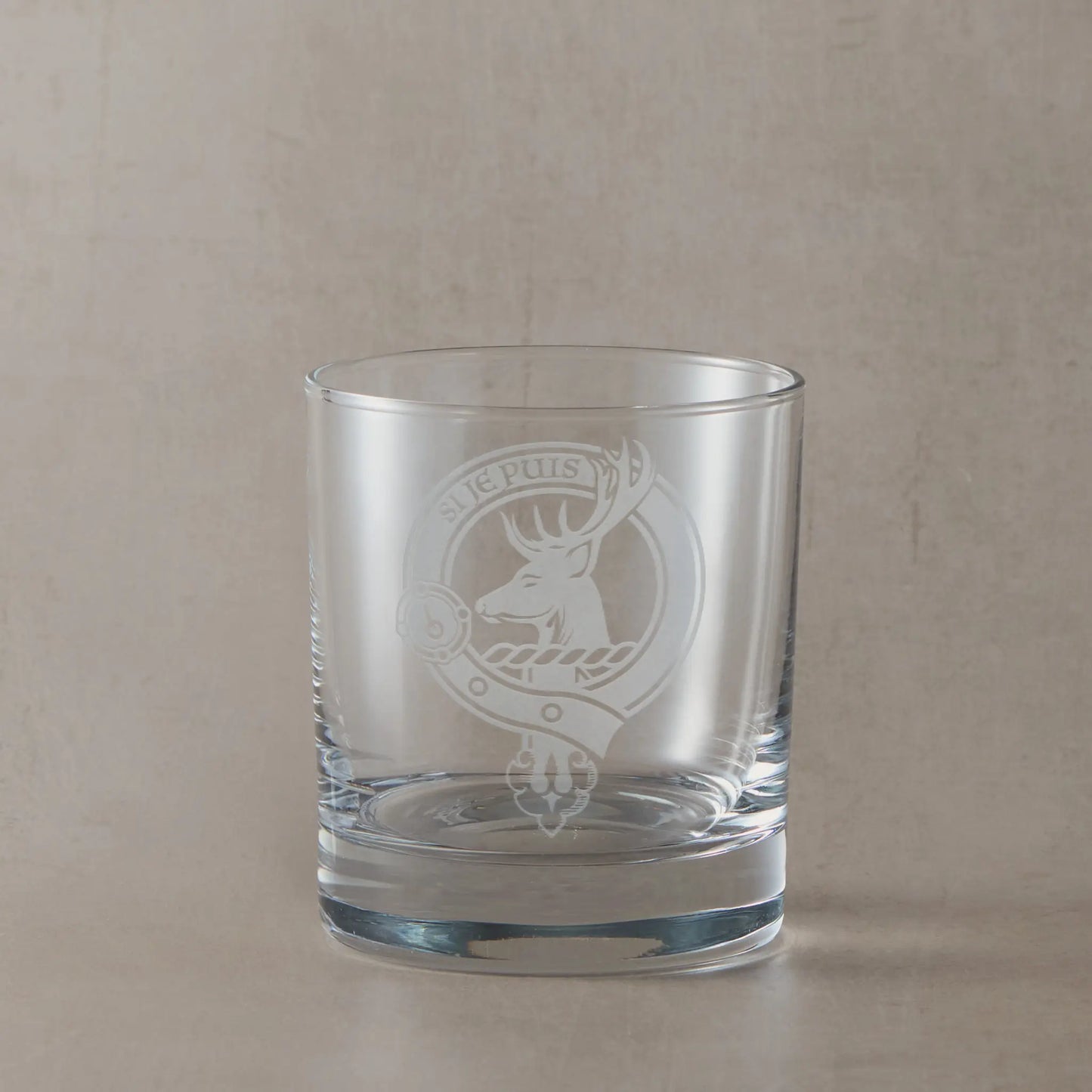 Engraved whisky glass featuring a Scottish clan crest with belt and buckle motif, sandblasted on a clear glass tumbler against a neutral beige studio background