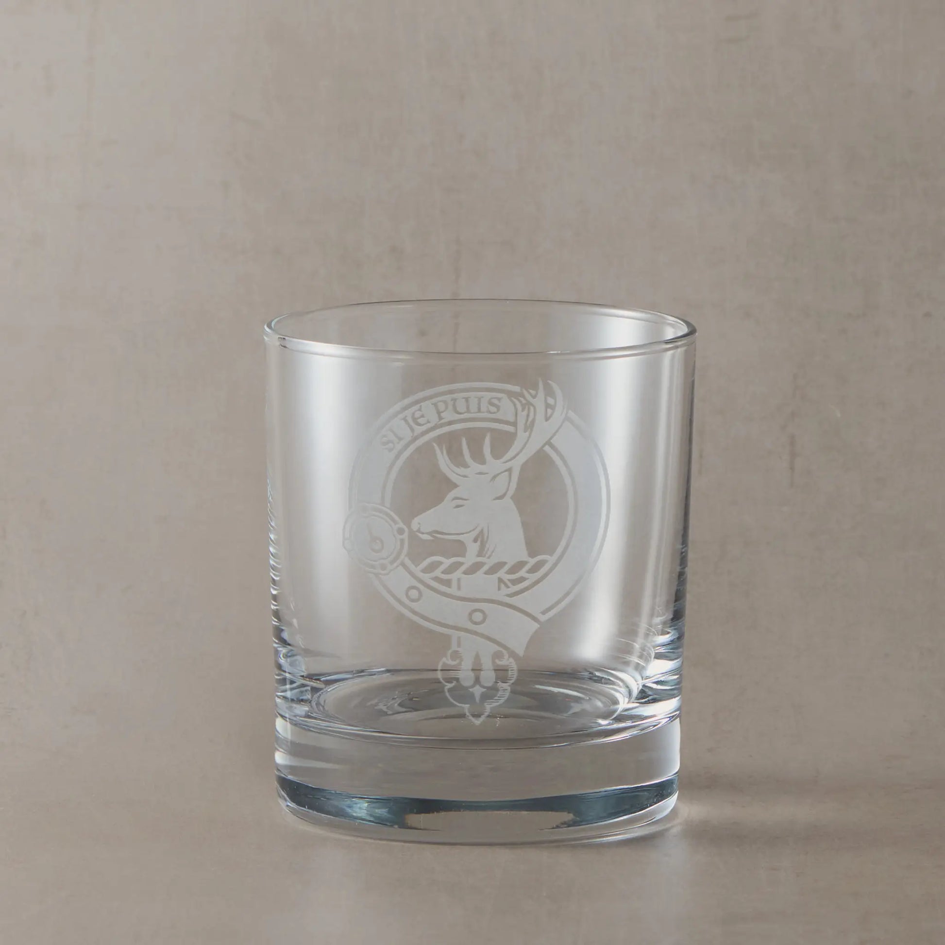Engraved whisky glass featuring a Scottish clan crest with belt and buckle motif, sandblasted on a clear glass tumbler against a neutral beige studio background