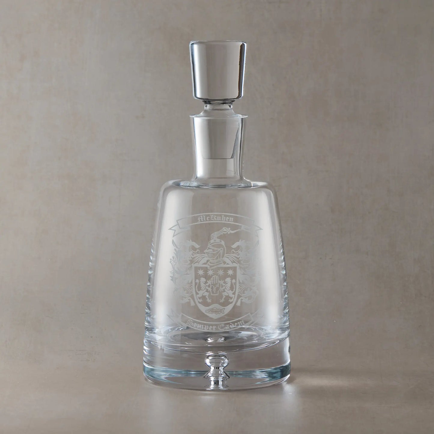 Handmade engraved spirit decanter featuring a heraldic coat of arms with central shield, ribbon banners for motto and surname, and a solid bubble base, set on a clear Atelier decanter against a neutral beige studio background