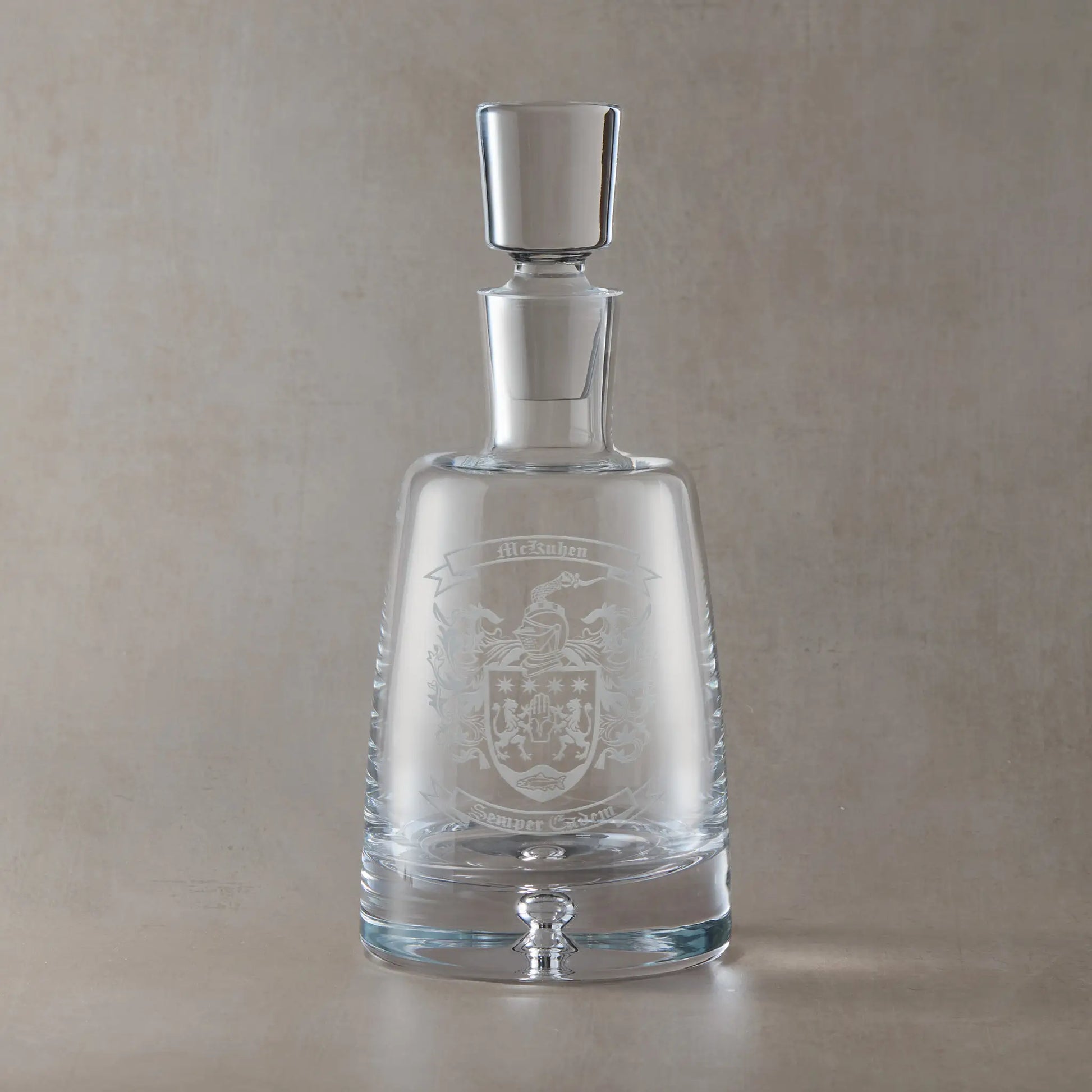 Handmade engraved spirit decanter featuring a heraldic coat of arms with central shield, ribbon banners for motto and surname, and a solid bubble base, set on a clear Atelier decanter against a neutral beige studio background