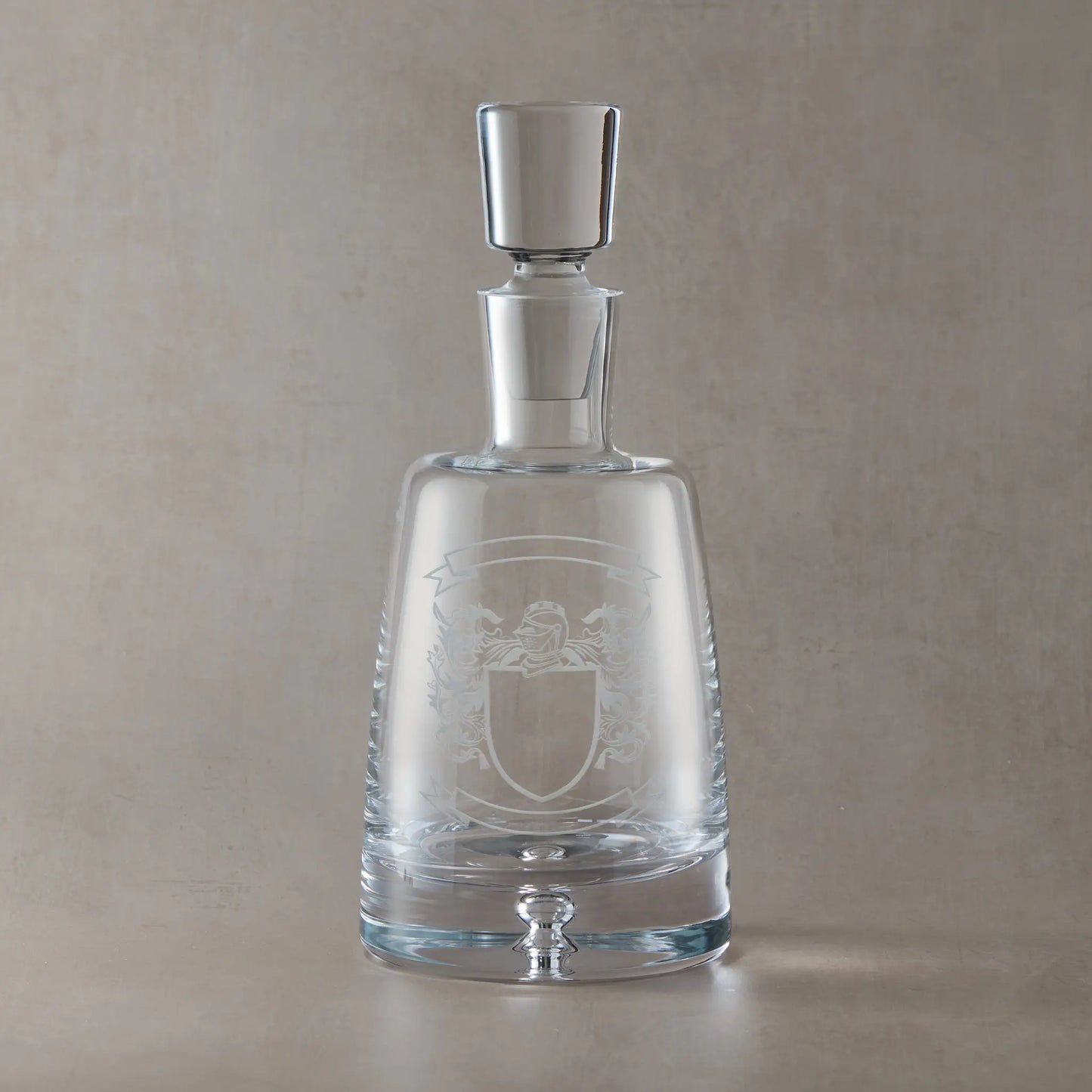 Engraved spirit decanter featuring a heraldic coat of arms with mantle and central shield, left blank for your own design, with ribbon banners for motto and surname, set on a clear Atelier decanter against a neutral beige studio background
