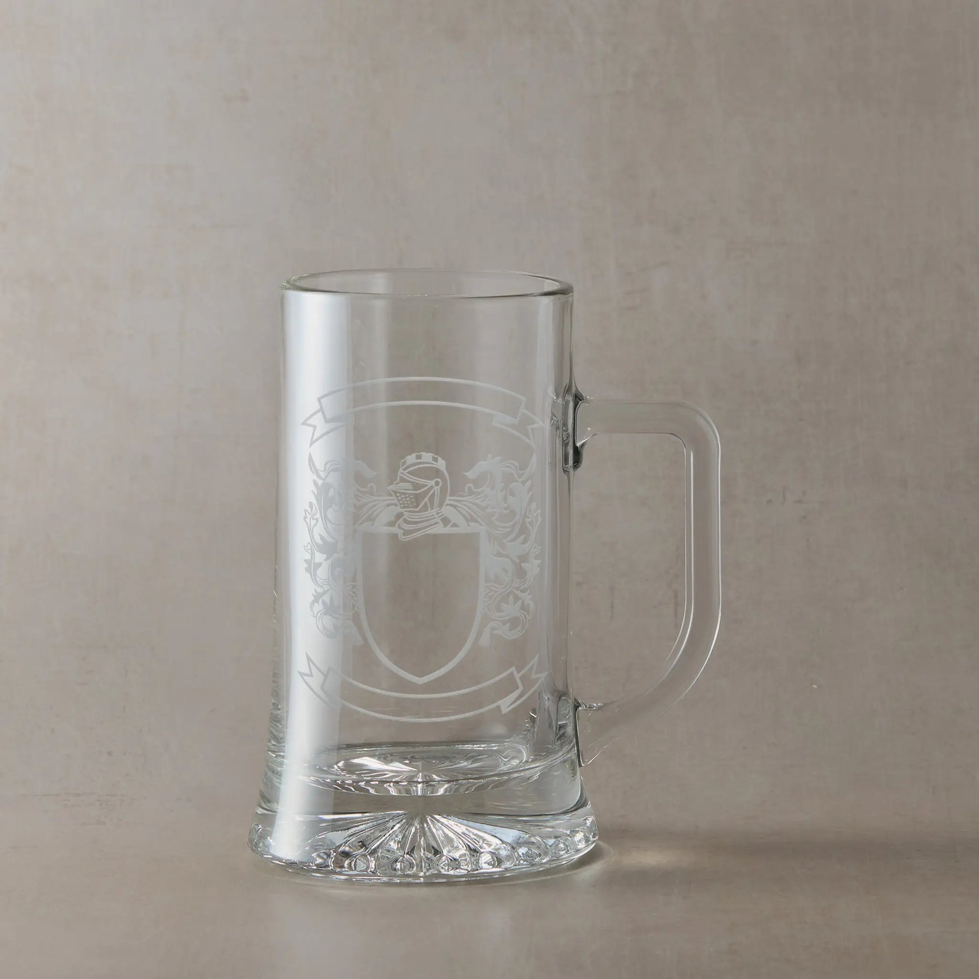 Engraved glass tankard featuring a heraldic coat of arms with a central shield left clear for your own design, handle on the right, set against a neutral beige studio background