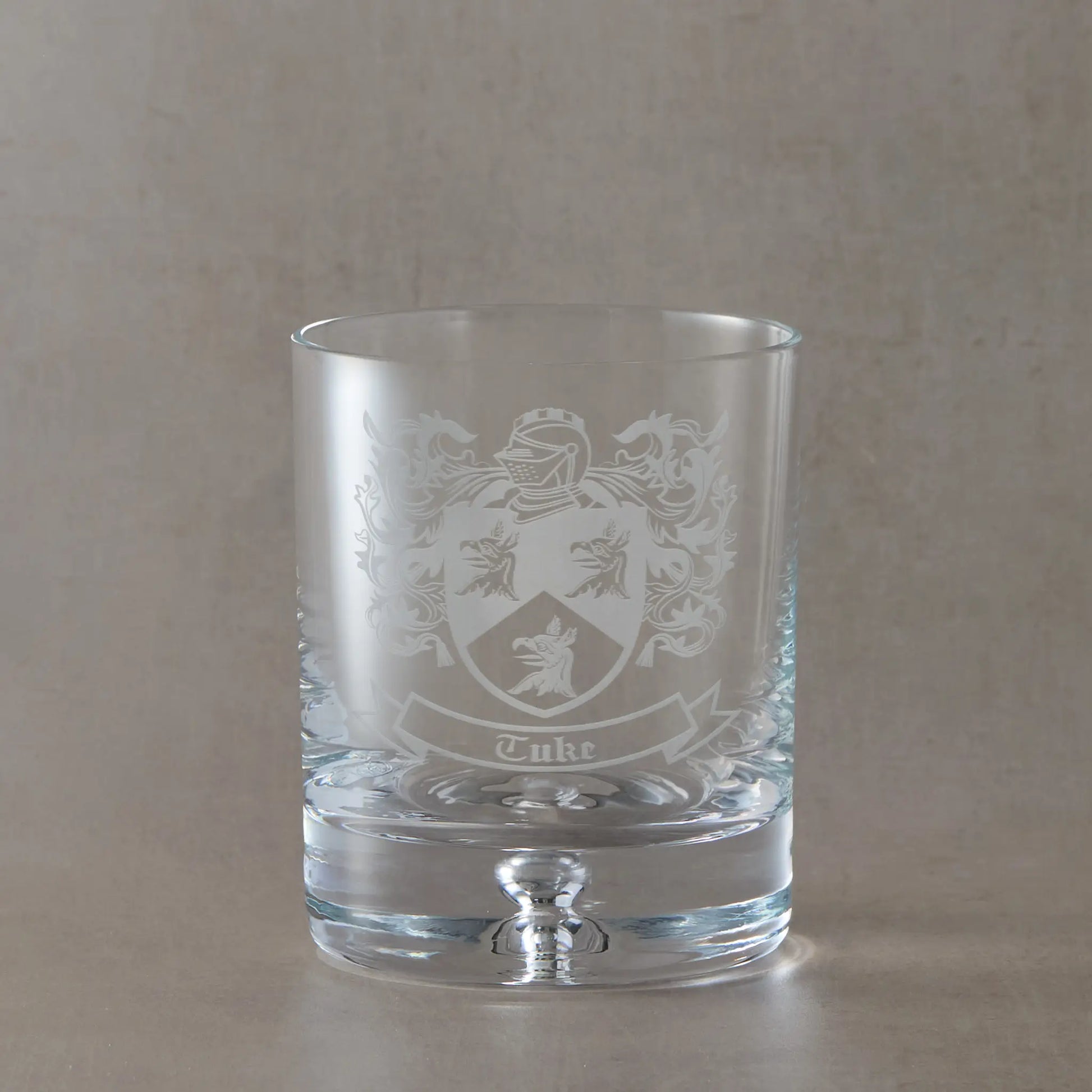 Whisky Engraved whisky glass featuring a heraldic coat of arms with mantel, shield and surname, sandblasted on a clear Atelier whisky glass against a neutral beige studio background