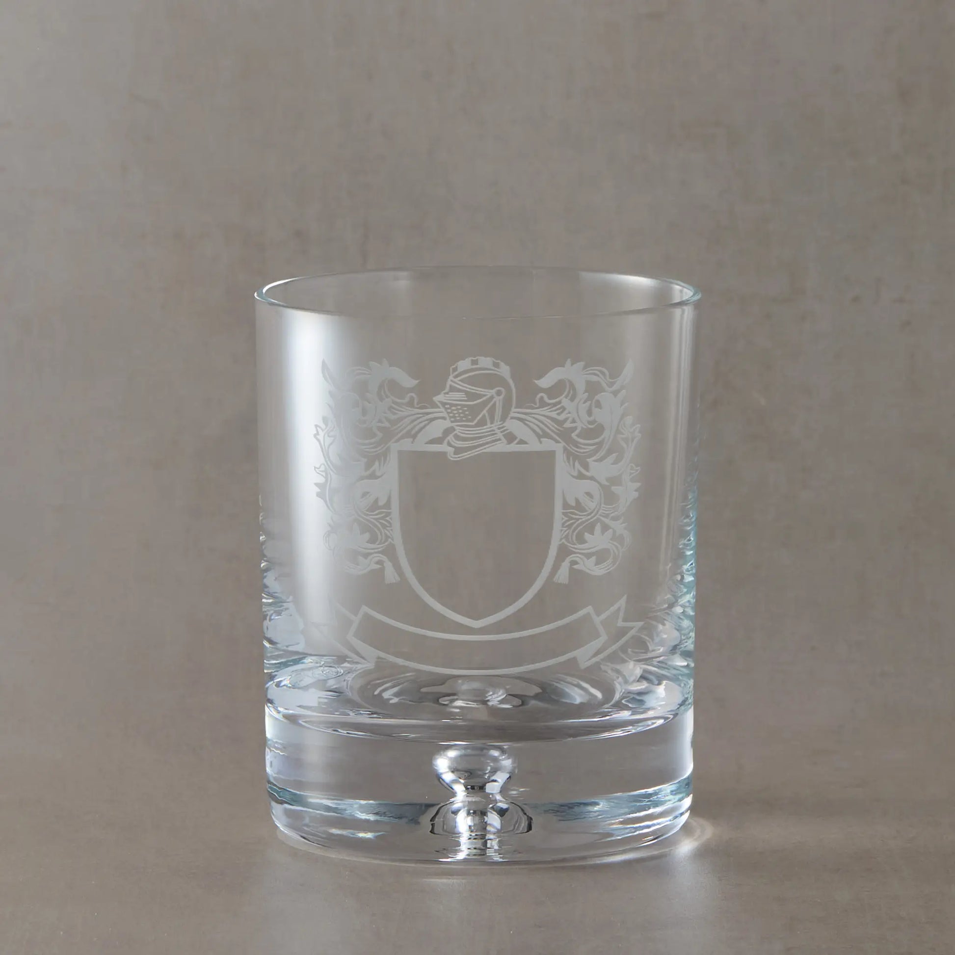 Engraved whisky glass with heraldic coat of arms design on a bubble base tumbler, shown on a neutral beige studio background
