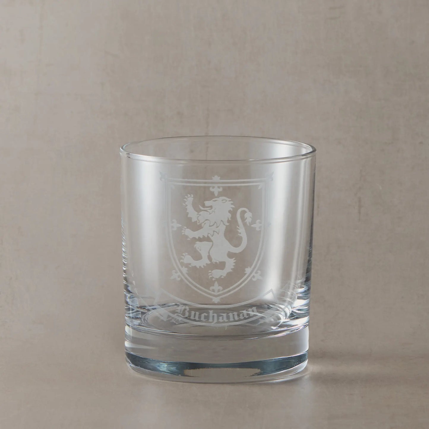 Engraved whisky glass featuring a heraldic coat of arms with family surname beneath, sandblasted on a clear glass tumbler against a neutral beige studio background