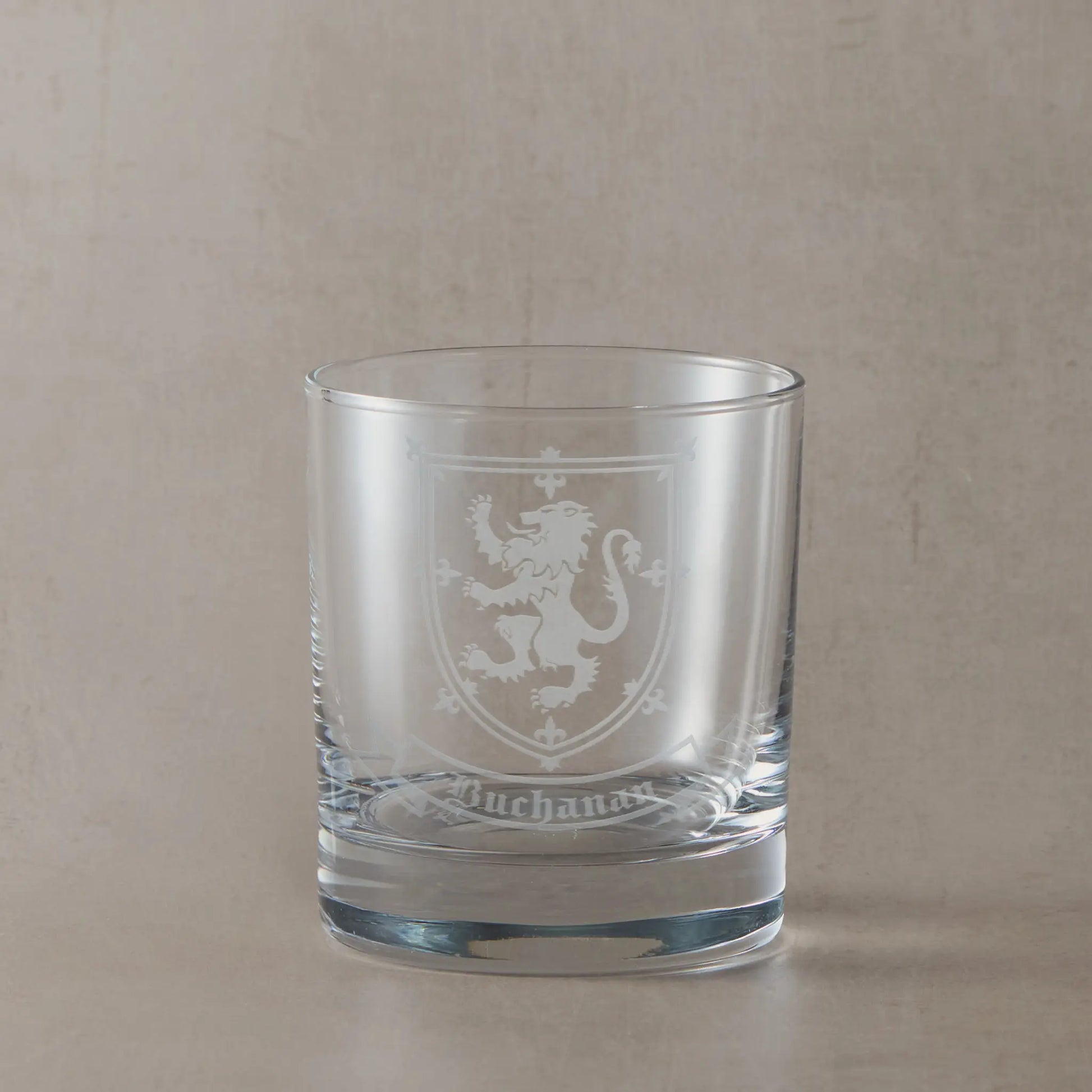 Engraved whisky glass featuring a heraldic coat of arms with family surname beneath, sandblasted on a clear glass tumbler against a neutral beige studio background