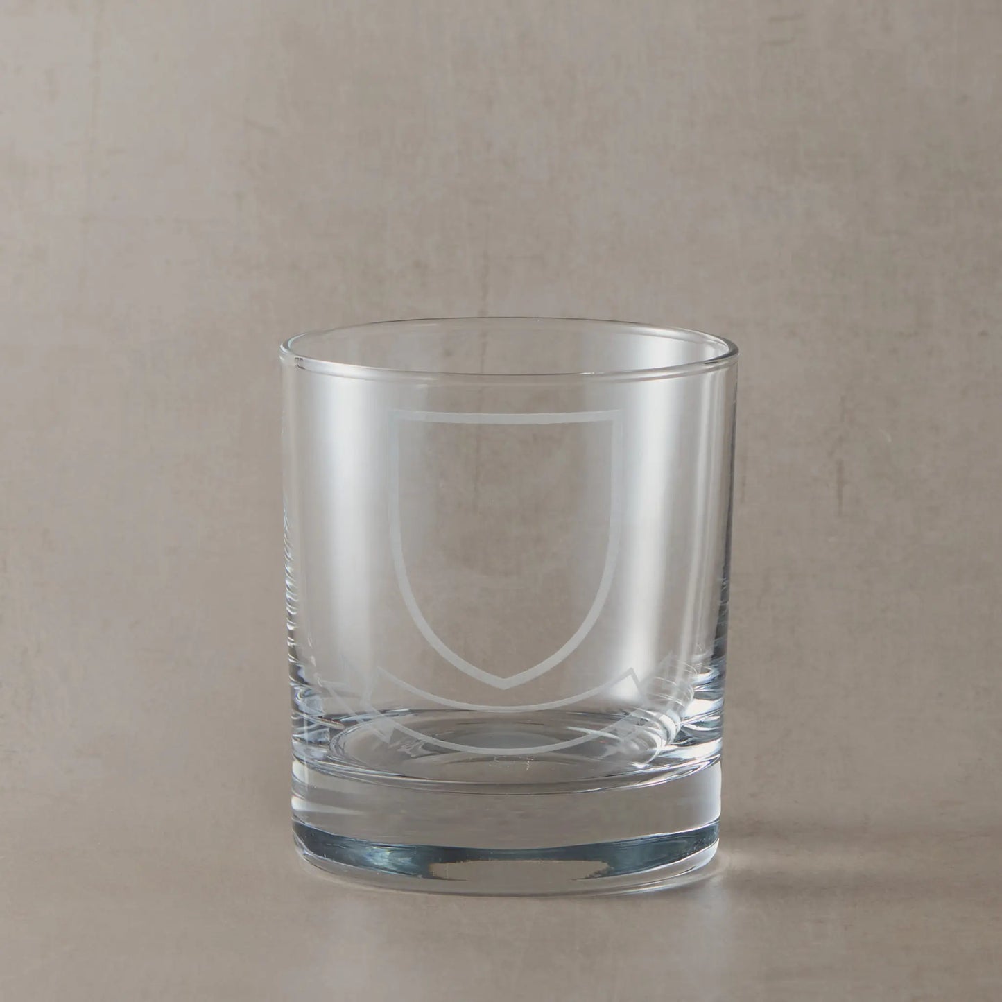 Engraved whisky glass featuring a heraldic coat of arms with shield and ribbon banners, left blank for your own family design, on a clear glass tumbler against a neutral beige studio background