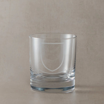 Engraved whisky glass featuring a heraldic coat of arms with shield and ribbon banners, left blank for your own family design, on a clear glass tumbler against a neutral beige studio background