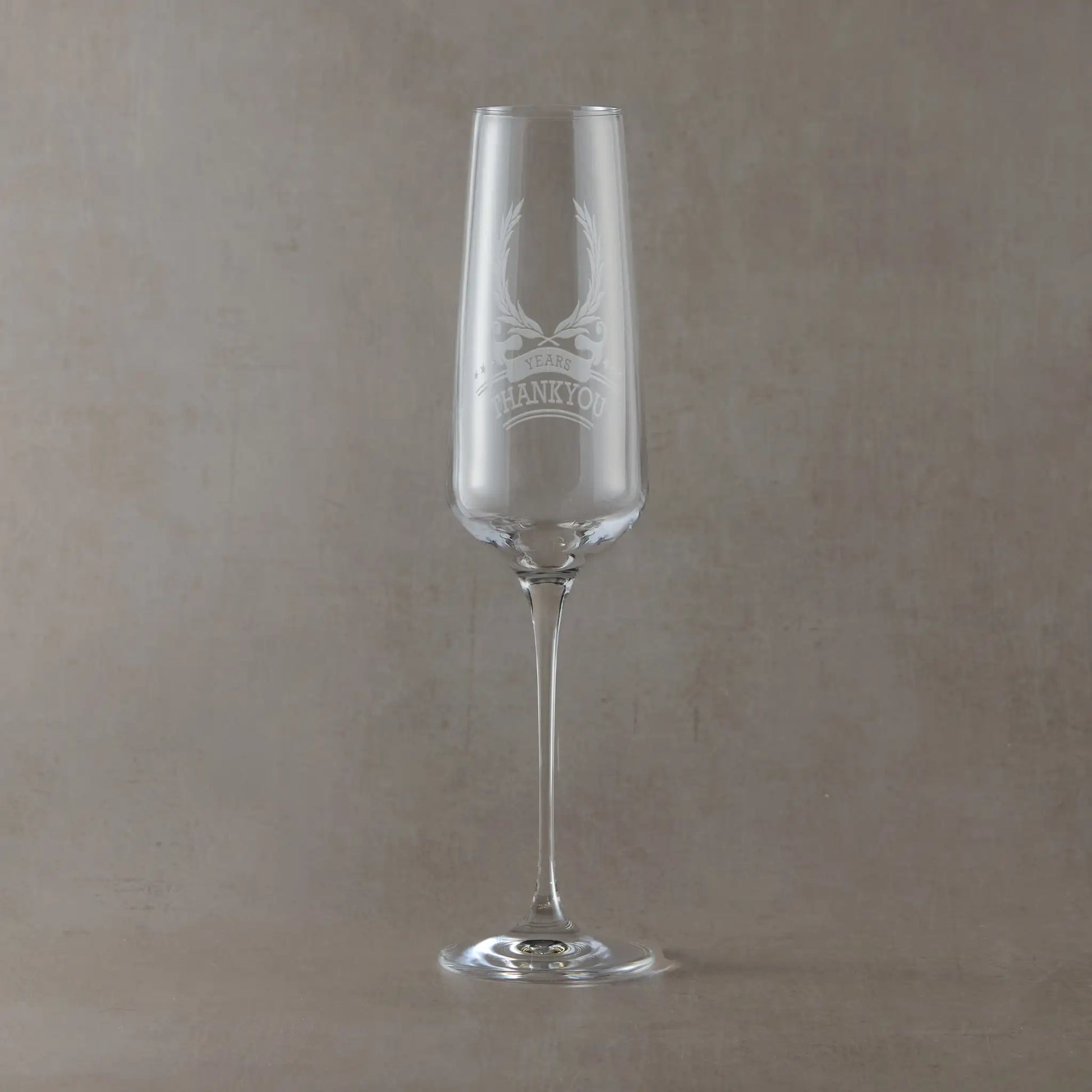 Champagne flute engraved with laurel wreath, ribbon anniversary design and thankyou photographed on a beige background