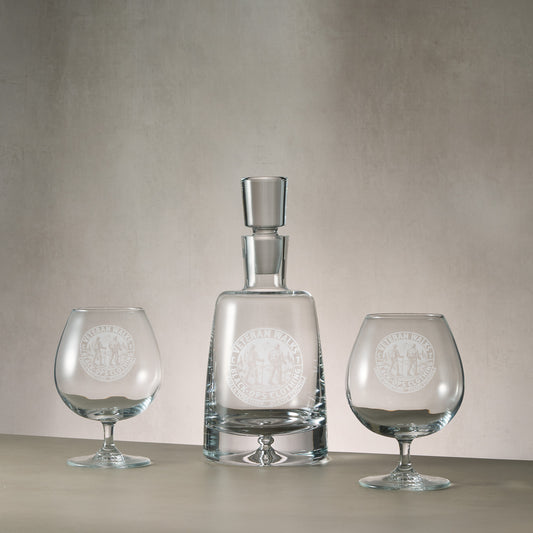 A clear glass crystal decanter featuring a company logo and two matching engraved brandy glasses.