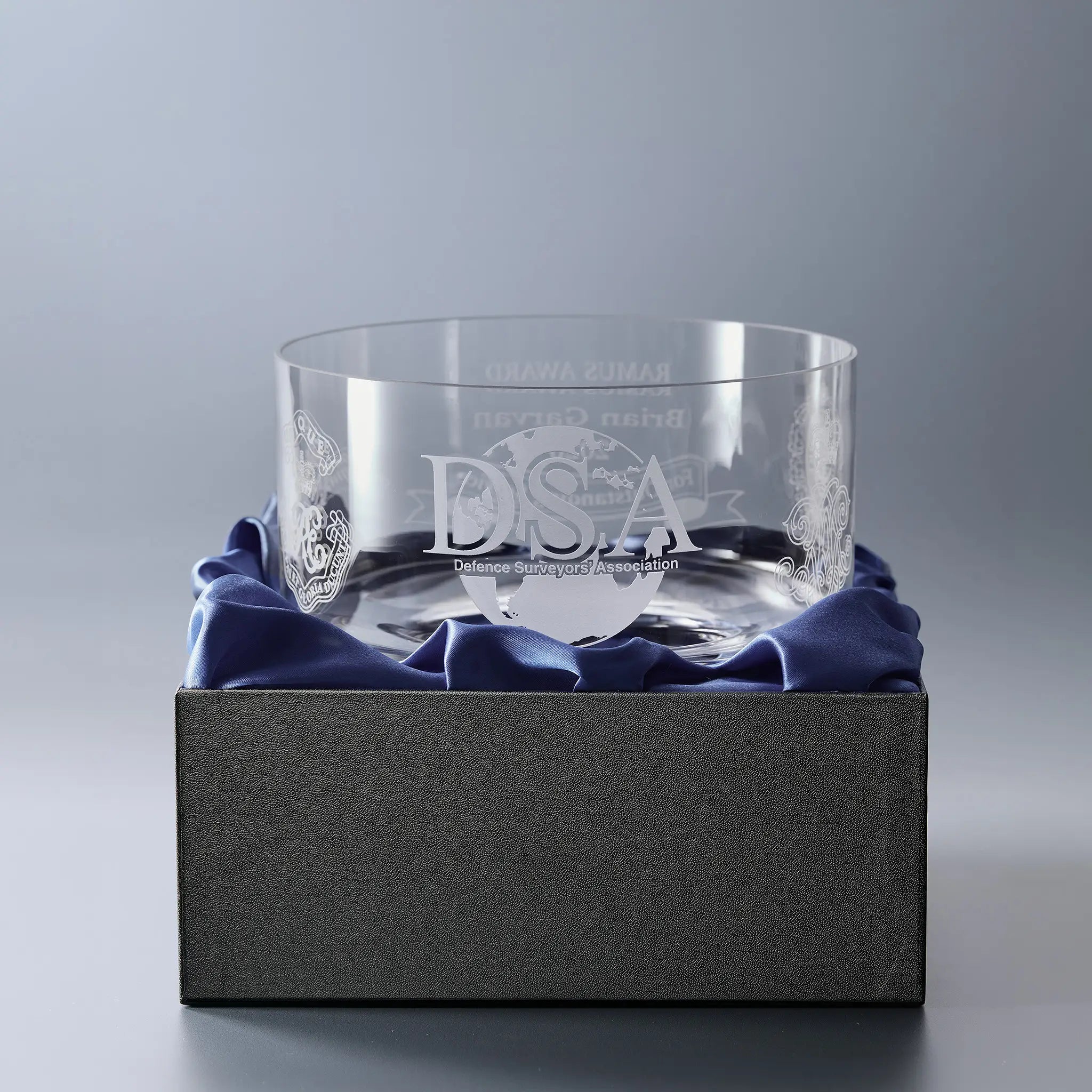 A clear glass bowl engraved with 'DSA' logo and text, displayed inside a dark gray box lined with blue satin fabric.