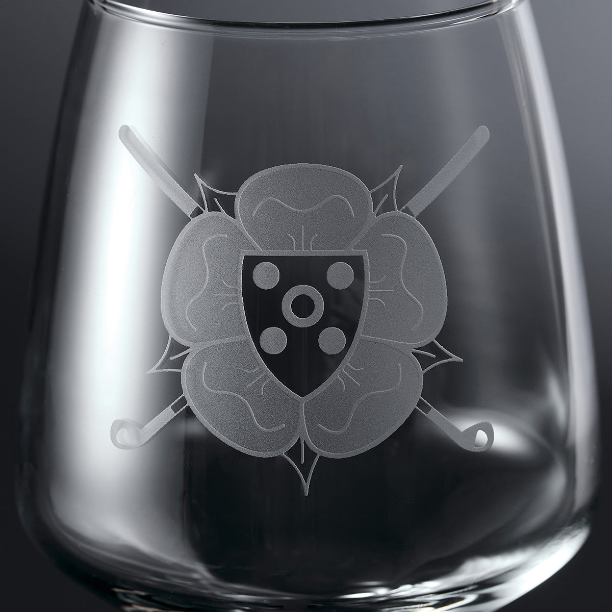 A close-up of a clear stemless wine glass with an etched golf club shield and floral design on the front.