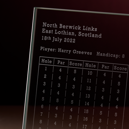 A close-up of an engraved glass golf scorecard displaying the course, date, players name ,hole, par and score
