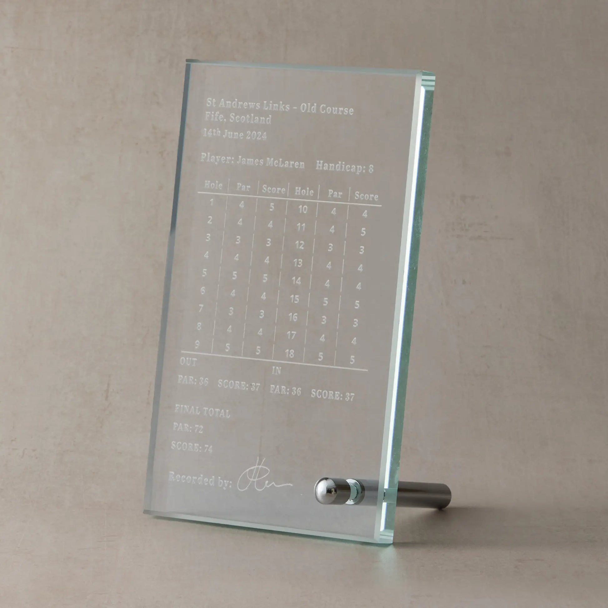 Engraved glass display plaque showing a personalised golf scorecard, photographed upright on a neutral stone-toned background.