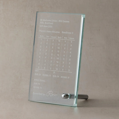 Engraved glass display plaque showing a personalised golf scorecard, photographed upright on a neutral stone-toned background.