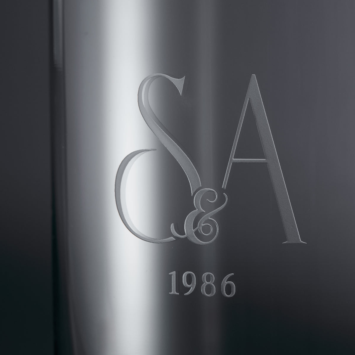 Close-up of a sandblast engraved vase featuring a monogram of 'S&A' with the year '1986' below it, set against a dark gray gradient background.