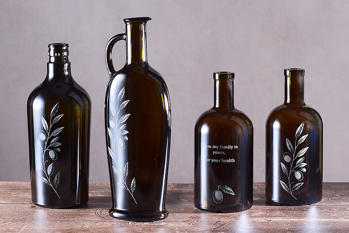 Four dark amber glass bottles with engraved olive branch designs and one with etched text.