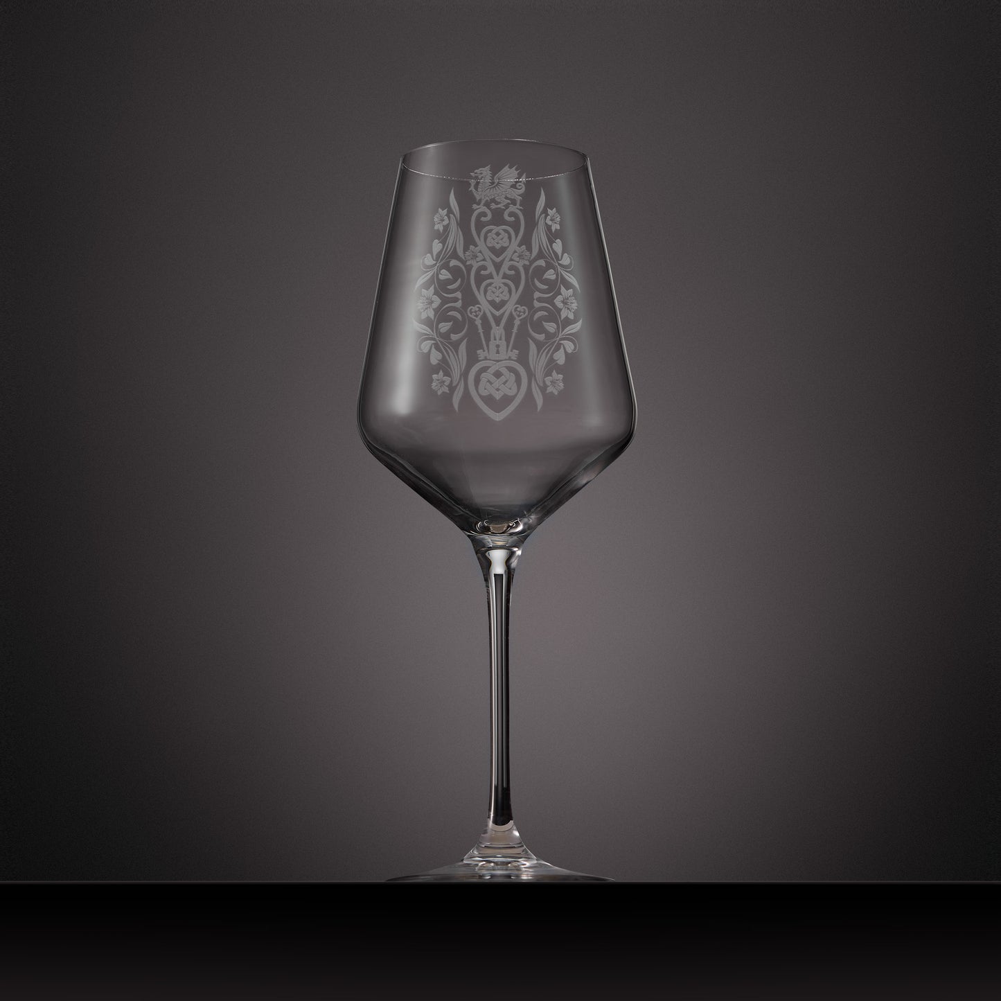 A clear glass wine glass with an etched ornate Welsh lovespoon design on the bowl.