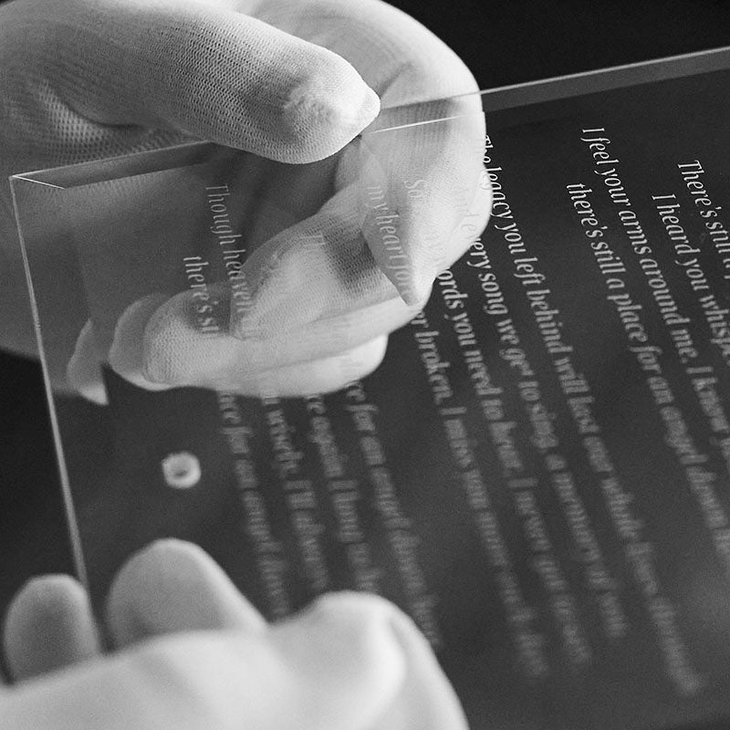 A close-up of a person wearing white gloves holding a transparent glass plate with engraved with a personalised poem