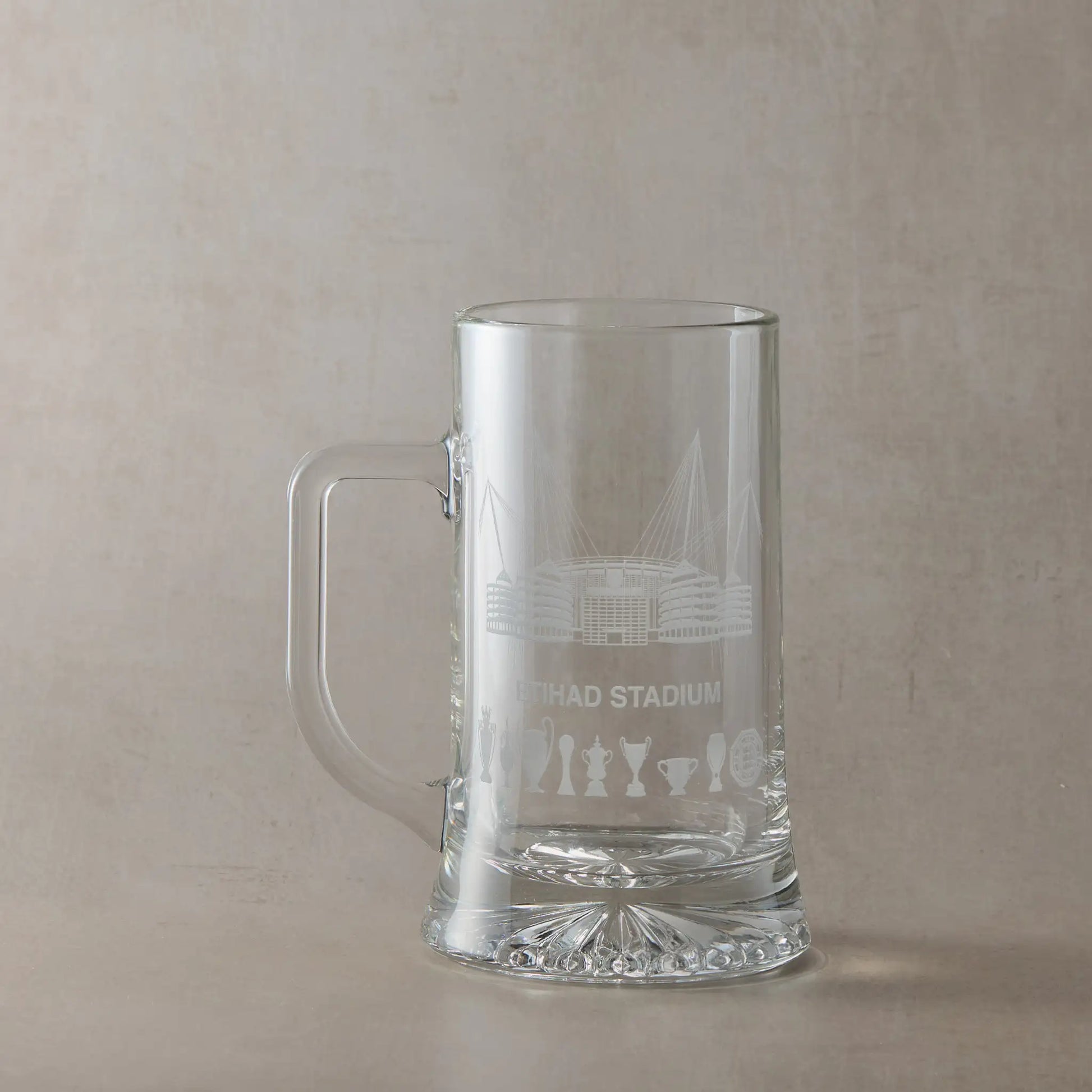 Clear glass tankard engraved with a line-art interpretation of the Etihad Stadium and trophy silhouettes, photographed on a beige background.