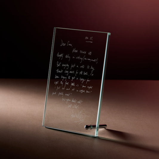Glass display plaque with engraved handwritten letter, photographed on a dark, softly lit background