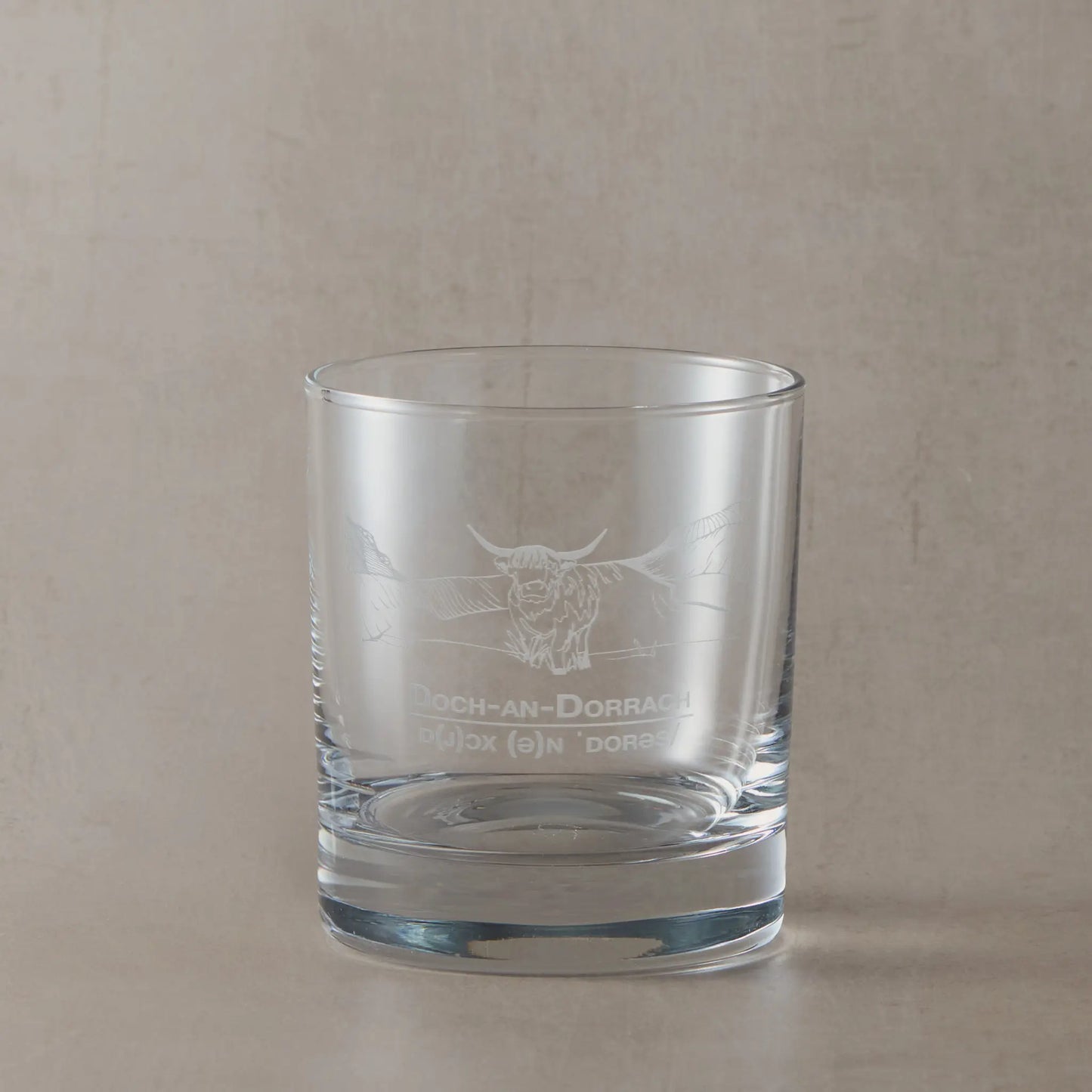 Clear glass tumbler with engraved highland cow and Gaelic text design on a beige background