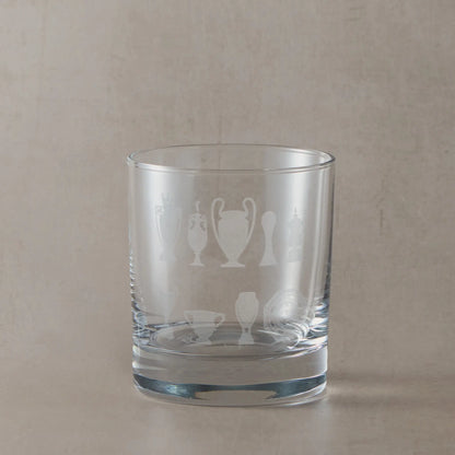 Whisky glass engraved with nine trophy silhouettes photographed on a beige background