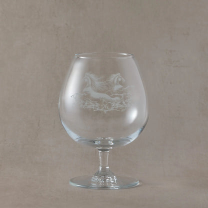 Clear brandy glass engraved with two galloping kelpies, captured mid-motion with flowing manes and water beneath, photographed on a soft neutral beige background