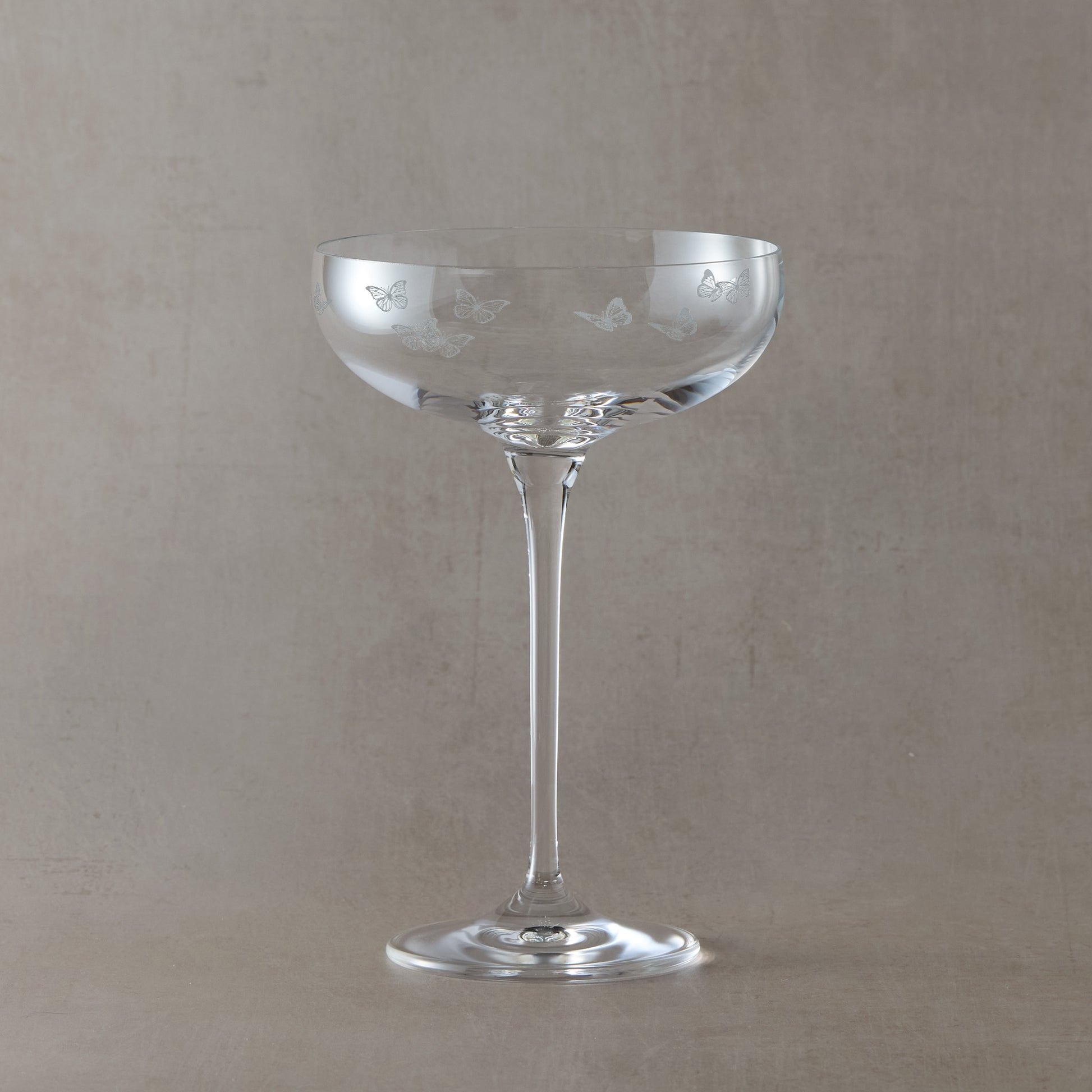 Clear glass champagne coupe with butterfly etching on a beige background
