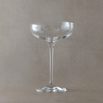 Clear glass champagne coupe with butterfly etching on a beige background
