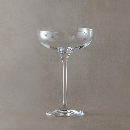 Clear glass champagne coupe with butterfly etching on a beige background