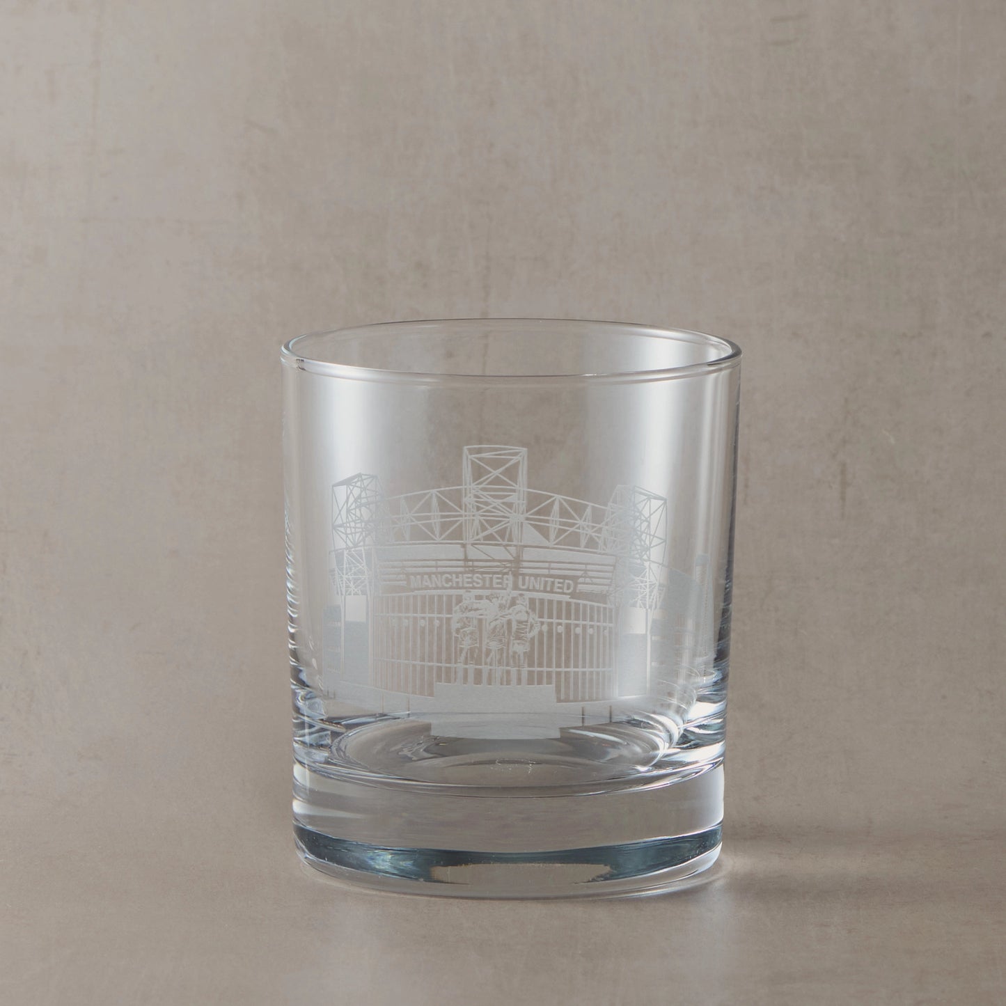 Whisky glass engraved with the facade of Old Trafford stadium, on a beige backround