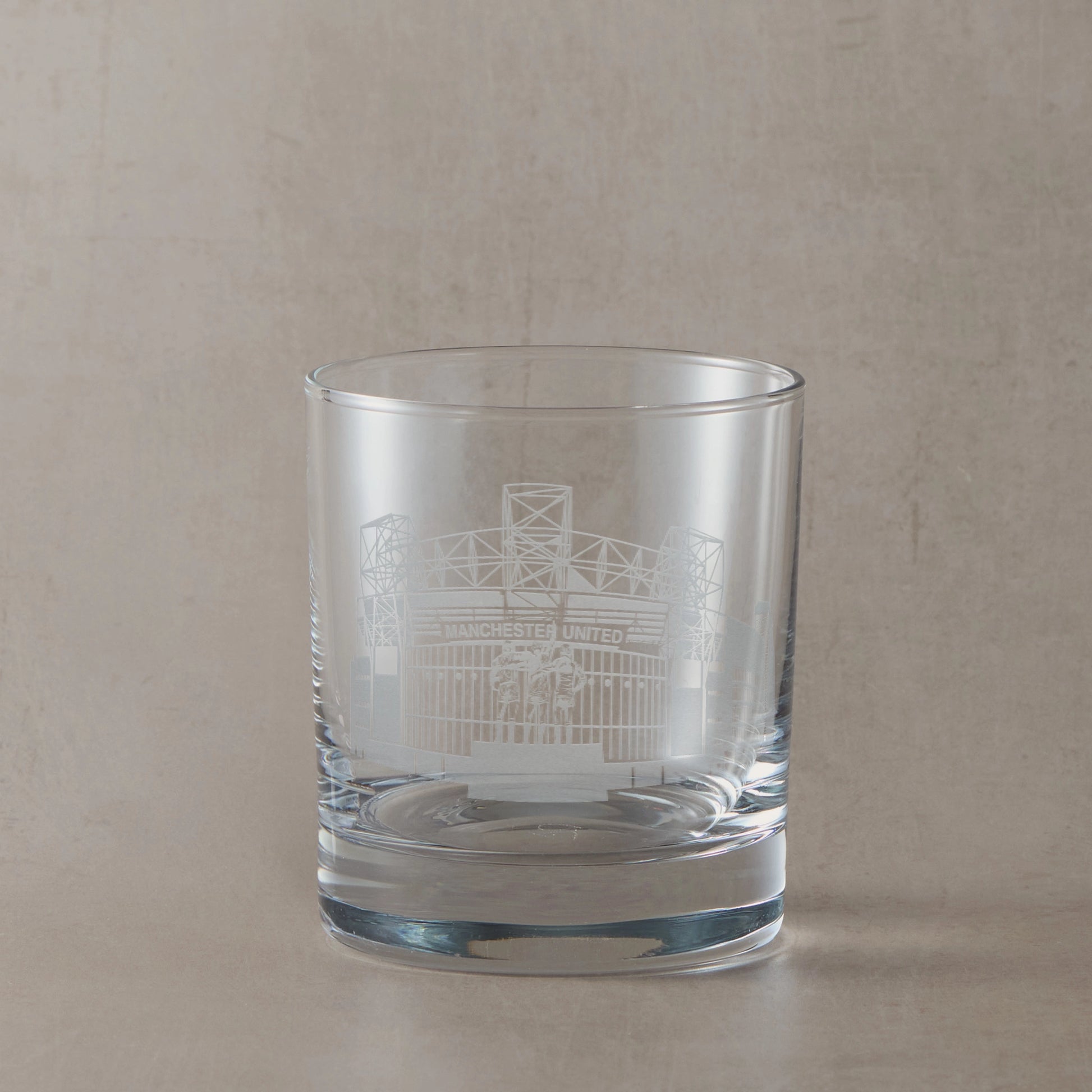 Whisky glass engraved with the facade of Old Trafford stadium, on a beige backround