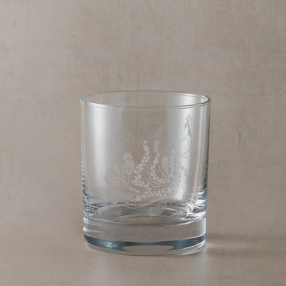 Engraved peacock on a whisky glass placed on a wooden surface with a bottle of Daftmill whisky in the background.