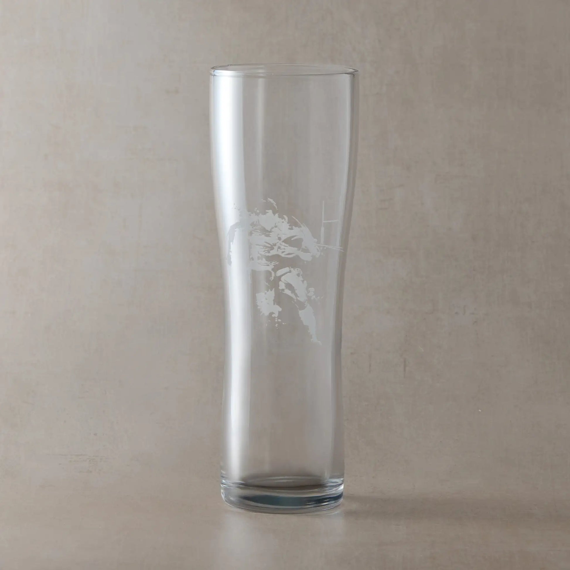 Tall pint glass engraved with a rugby player and ball, photographed on a beige background
