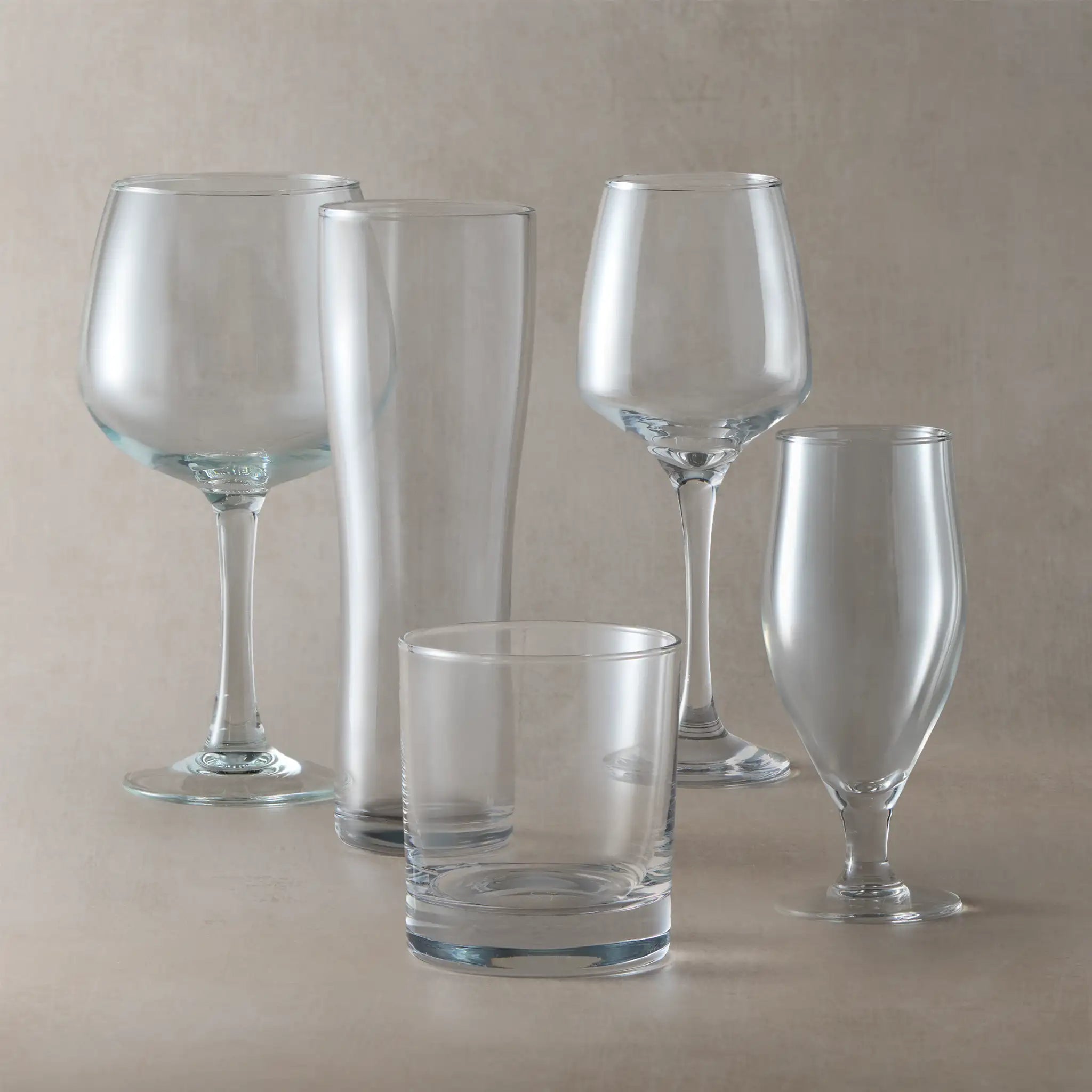 Set of clear glassware including wine glasses and a tumbler on a beige background